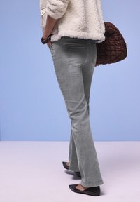 Personne portant un jean bootcut gris, des ballerines pointues noires, une veste en polaire crème et tenant un sac matelassé marron sur un fond bleu.