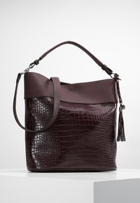 Sac fourre-tout bordeaux avec un motif texturé de crocodile sur le corps, dessus en cuir lisse et un accent de gland amovible. Bretelle réglable.