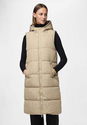 Beige hette puffer vest med glidelås, store lommer og quiltet design. Brukt over en svart ribbestrikket langermet topp.