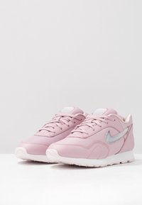 Vaaleanpunaiset tennarit, jotka on valmistettu synteettisestä materiaalista. Ne ovat verkkomaiset päältä, sisältävät hopeanvärisen Nike-logon ja valkoisen kumipohjan.