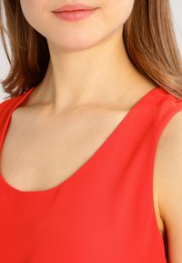Primo piano della parte inferiore del viso di una donna, collo e spalle, che indossa una maglietta rossa senza maniche con lunghi capelli castani visibili.