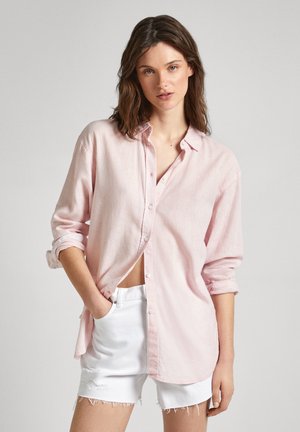 Camicia button-up rosa chiaro con maniche arrotolate, realizzata in un tessuto morbido, indossata sopra pantaloni corti di jeans bianchi con bordi sfilacciati.