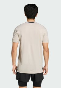 Jasno beżowa sportowa koszulka z krótkim rękawem, z zaokrąglonym dołem i czarnym panelem na karku. Zawiera branding "AEROREADY" na dole.