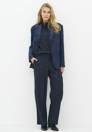 Jeune femme aux longs cheveux blonds portant un blazer bleu marine, une chemise bleu foncé boutonnée, un pantalon à jambes larges et des bottines noires, se tenant contre un mur blanc.