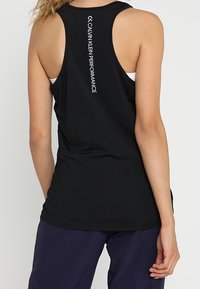 Frau in schwarzem ärmellosem Sportoberteil mit dem Aufdruck "Calvin Klein Performance" auf dem Rücken, kombiniert mit dunkelblauen Hosen.
