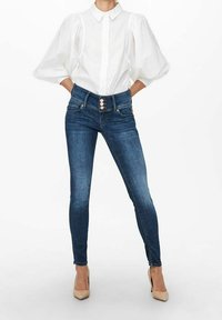 Mörkblå, mörk denim skinny jeans med hög midja, dekorativ knappstängning och fickor, kombinerade med en vit skjorta med ballongärmar.