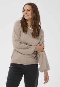 Donna con lunghi capelli castani che indossa un maglione beige con maniche a palloncino e jeans neri, sorridente e che guarda di lato su uno sfondo bianco.