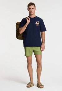 Tricou bleu marin cu design grafic, pantaloni scurți verzi și sandale maro. Modelul ține o geantă verde măsliniu, stând pe un fundal simplu.