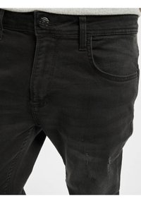 Detailní záběr černých džínsů z denimu s předními kapsami, zapínáním na knoflíky, poutky na pásek a jemným opotřebením na stehně.