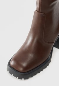 Botte en cuir marron avec un bout carré, semelle en caoutchouc noir épais et talon carré sur fond blanc.