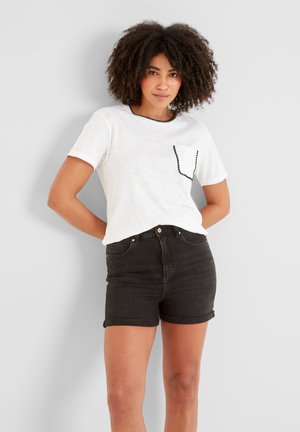 Young woman with curly hair wears white short-sleeve top with black trim and black rolled-up denim shorts, standing against a plain wall.