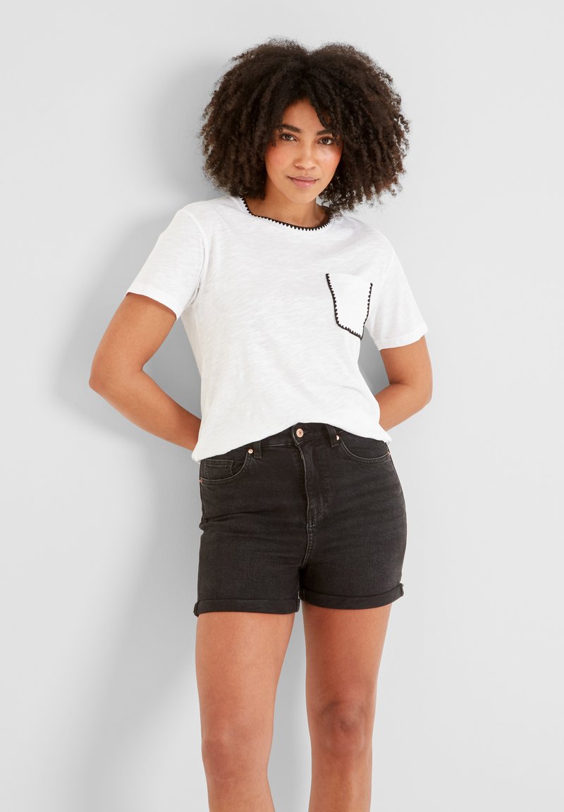Young woman with curly hair wears white short-sleeve top with black trim and black rolled-up denim shorts, standing against a plain wall.