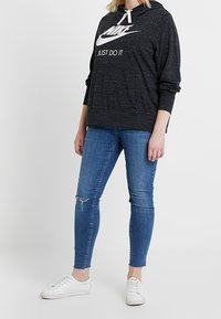 Sudadera negra con el logo blanco de "Nike Just Do It", combinada con jeans ajustados azules y zapatillas blancas. La capucha tiene cordones y tiene un ajuste casual.