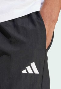 Calções atléticos pretos com um acabamento texturizado, apresentando o logótipo da Adidas bordado a branco e bolsos laterais. O tecido parece leve e respirável.