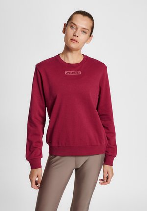 Frau steht und trägt ein kastanienbraunes Sweatshirt mit „Hummel“-Logo und beige Leggings vor einem einfarbigen hellen Hintergrund.