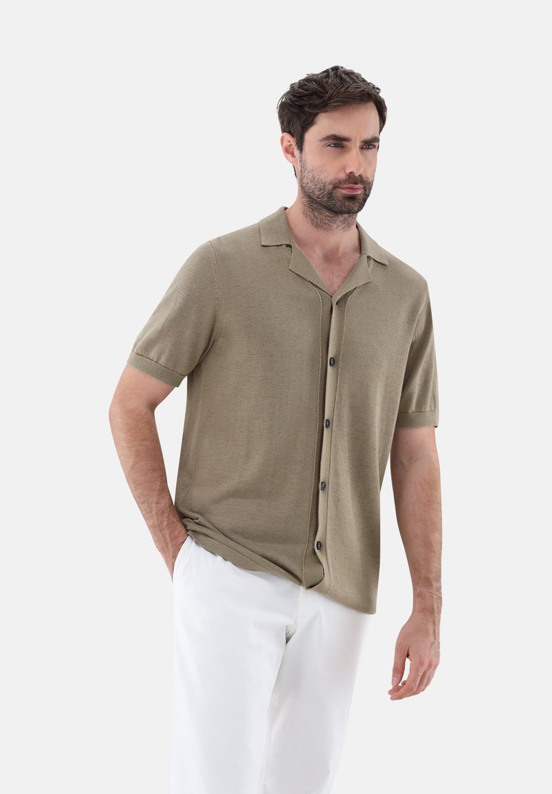 Camicia a maniche corte di colore verde oliva, con colletto, dalla texture liscia e cinque bottoni scuri lungo la parte frontale, abbinata a pantaloni bianchi.