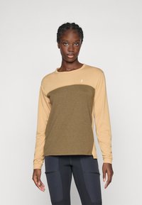 Vrouw met vlechten, gekleed in een beige en olijfgroene blouse met lange mouwen en donkere leggings met ritszakken, tegen een effen achtergrond.