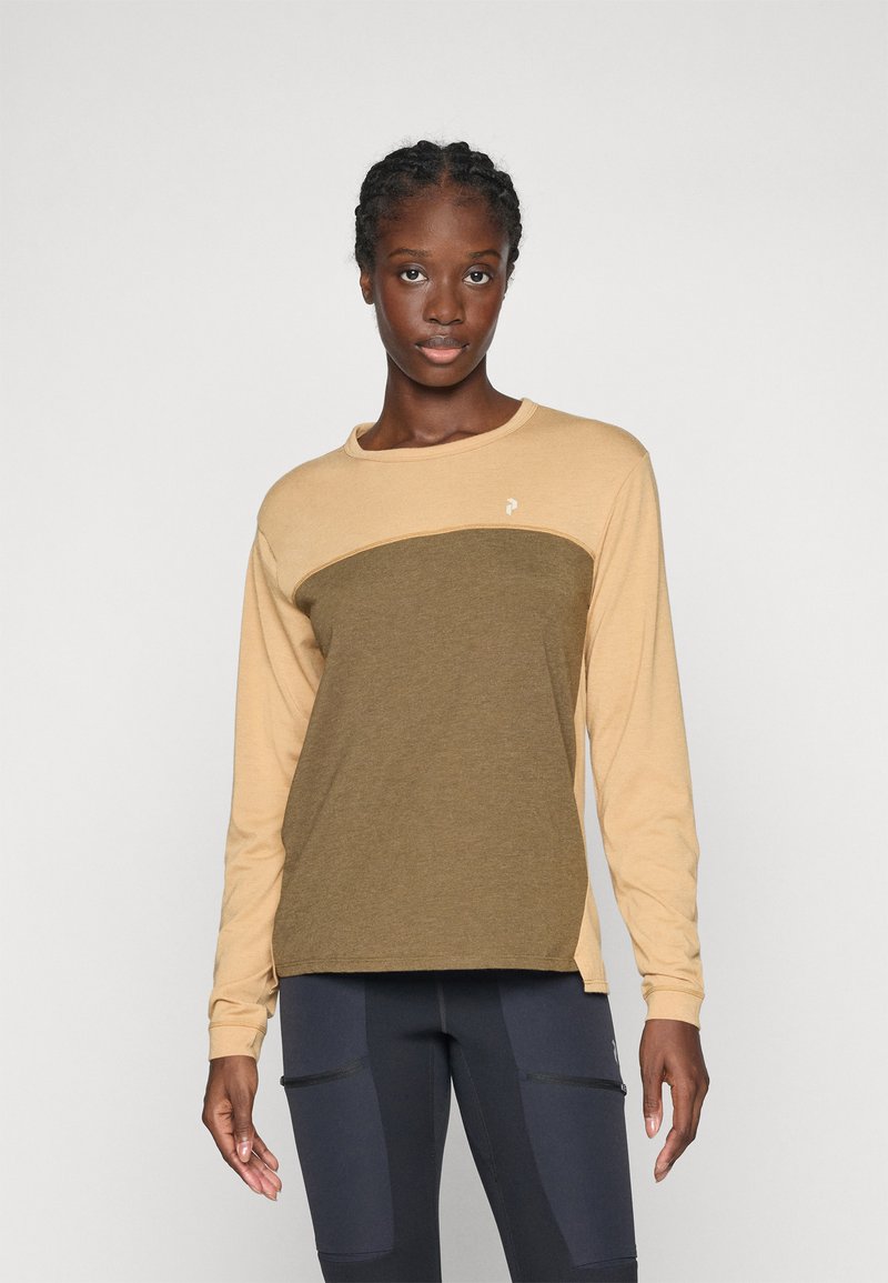 Vrouw met vlechten, gekleed in een beige en olijfgroene blouse met lange mouwen en donkere leggings met ritszakken, tegen een effen achtergrond.