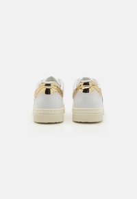 Sneakers bianche con un accento dorato lucido sul tallone, suola di crema testurizzata e linguetta bianca imbottita per il comfort.