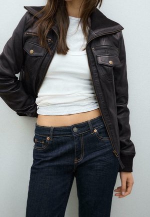 Frau trägt eine dunkelbraune Lederjacke über einem weißen kurzen Oberteil und dunklen blauen Jeans, steht vor einem einfarbigen hellen Hintergrund.