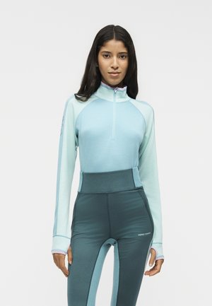 Joven mujer con cabello largo y oscuro que lleva una camiseta azul claro con cremallera y leggings azul oscuro de talle alto con agujeros para los pulgares.