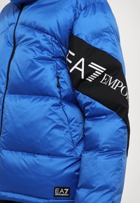 Plava puffer jakna s sjajnim završetkom, crni tkaninski akcent na rukavu sa bijelim logotipima i zippered džepovima na prednjoj strani.