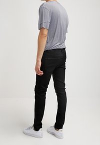 Person som bär en randig kortärmad skjorta, svarta slim-fit byxor och vita sneakers, står mot en enfärgad ljus bakgrund.