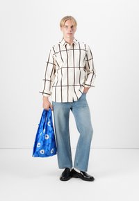 Camisa xadrez em tons de creme e castanho com botões, calças flare de ganga azul claro e uma bolsa de mão azul com estampado floral. Sapatos pretos tipo slip-on.