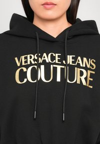 Svart hettegensere med gulltekst "VERSACE JEANS COUTURE." Har en justerbar hette og er laget av et mykt, teksturert stoff.