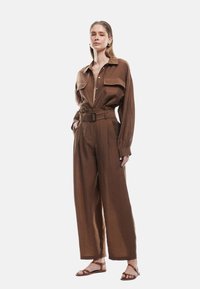 Brun linned jumpsuit med lange ærmer, brystlommer, en bælteliv og brede bukser. Sandalerne har et stroppesystem.
