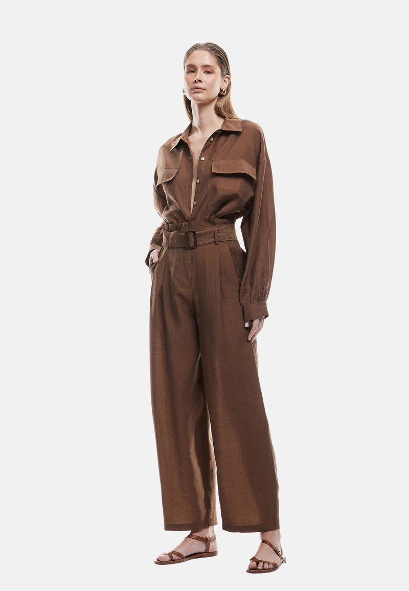 Brun linned jumpsuit med lange ærmer, brystlommer, en bælteliv og brede bukser. Sandalerne har et stroppesystem.