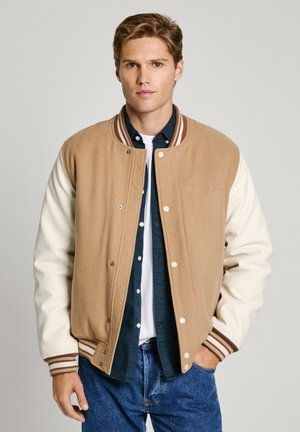 Brun og creme varsity-jakke med ribbede manchetter og krave, med trykknapper og broderet logo på brystet. Båret over en mørk skjorte.