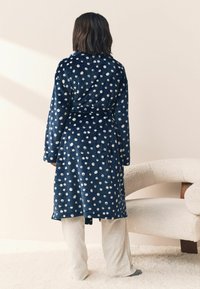 Asmuo, dėvintis tamsiai mėlyną chalato su taškeliais, surištą ties juosmeniu, stovintis ant kilimu dengto grindų šalia kreminės tekstūros lenkto fotelio.