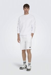 Joven de pie con un sudadera blanca de manga larga, pantalones cortos blancos con cordón, calcetines a rayas negras y blancas, y zapatillas blancas.