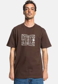 Camiseta de algodón marrón con mangas cortas. Presenta un diseño gráfico de logo blanco en el centro. Corte clásico y textura suave.