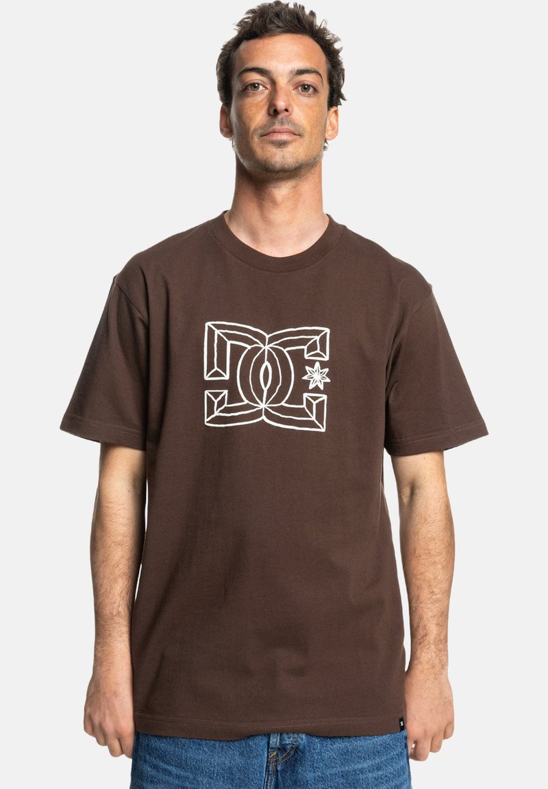 Camiseta de algodón marrón con mangas cortas. Presenta un diseño gráfico de logo blanco en el centro. Corte clásico y textura suave.