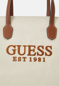 Beige canvas tote bag with brown leather handles and white stitching. Features large brown embroidered "GUESS" and "EST 1981" text.