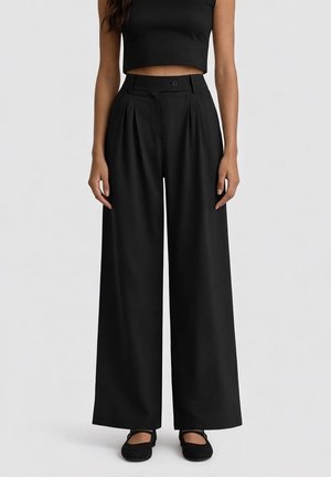 Donna con pantaloni neri a vita alta e gamba larga, top nero corto senza maniche e scarpe Mary Jane nere, in piedi davanti a uno sfondo neutro.