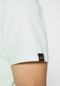 En persons överarm som bär en ljus mintgrön kortärmad skjorta med en svart etikett som har en guldstjärnelogo på ärmslutet.