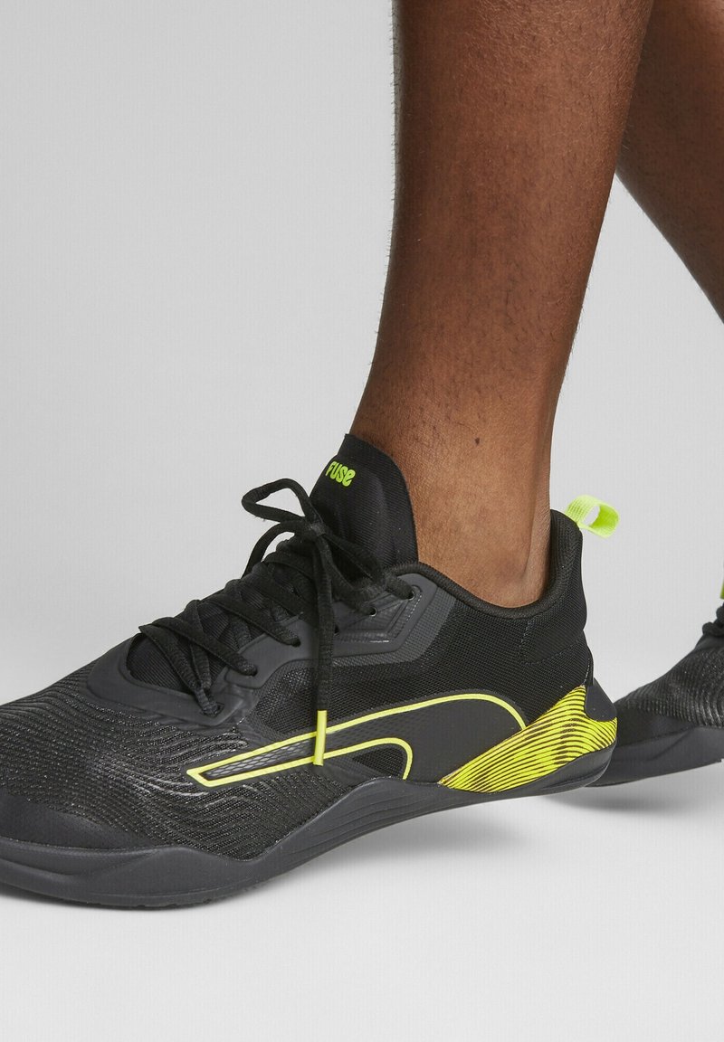 Chaussures de sport noires avec des touches jaunes portées pieds nus, montrant des lacets attachés et un design texturé sur un fond clair uni.