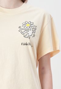 Light yellow cotton t-shirt featuring a graphic of a dancing figure and a daisy with a yellow center. Short sleeves and a relaxed fit.