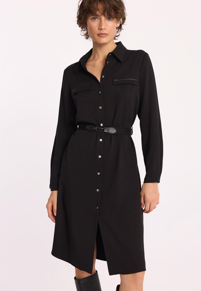 Czarna sukienka koszulowa z gładkiego materiału, z zapinanym przodem na guziki, dwoma kieszeniami na piersi oraz paskiem w talii. Długie rękawy dopełniają całość.