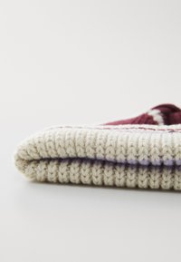Coperta lavorata a maglia con una base crema, caratterizzata da strisce marrone scuro e viola. La trama è morbida e spessa, evidenziando una qualità accogliente e fatta a mano.