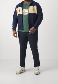 Felpa con zip color block navy e crema sopra una t-shirt verde, abbinata a pantaloni slim fit scuri e sneakers bianche con dettagli verdi.