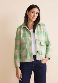 Giacca a quadri verde e beige con colletto, tasche frontali e chiusure a bottone, indossata sopra un top bianco. Tessuto liscio.