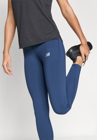 Tamnoplave atletske tajice izrađene od rastezljivog materijala, s prilagođenim dizajnom i malim bijelim logotipom na lijevom bedru. Glatka tekstura.