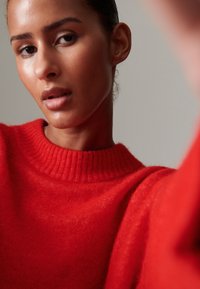 Röd ribbad stickad tröja med hög krage. Mjuk textur, avslappnad passform och en lätt oversize design. Närbild på halsringning och ansikte.