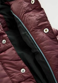 Maroon puffer jacketin sisäosa, jossa on silkkimäinen rakenne, hopeiset napit ja erottuva musta vuori reunoilla.