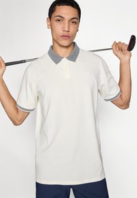 Κρεμ πόλο πουκάμισο με υφασμένο γκρι γιακά και μανσέτες. Διαθέτει κρυφό κόψιμο με κουμπιά και κεντημένο λογότυπο. Μοντέλο κρατάει ρόπαλο γκολφ.