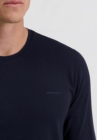 Mann in einem dunkelblauen Langarmshirt mit "Pierre Cardin"-Logo auf der Brust, von Hals bis zur oberen Körpermitte gezeigt.